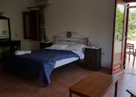 Hotel bedroom with a metal‑frame bed, navy bedspread, two nightstands, a dresser with a mirror, and open balcony doors to an outdoor seating area.