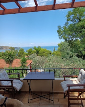 Balcony with a table and two cushioned chairs overlooking the blue sea and green trees beneath a wooden pergola.