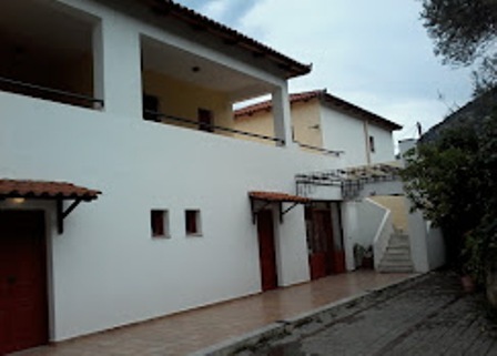 Two-story white townhouse-style building with a tiled walkway, brown doors, small square windows, and a right-side exterior staircase.
