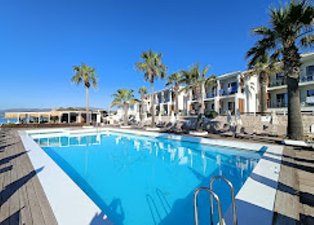 Outdoor resort pool surrounded by palm trees, lounge chairs, and white buildings on a sunny day.