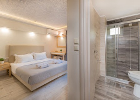 Bright hotel bedroom with a large bed dressed in white linens, two nightstands, a small desk, and an open doorway to a glass-enclosed bathroom visible on the right.