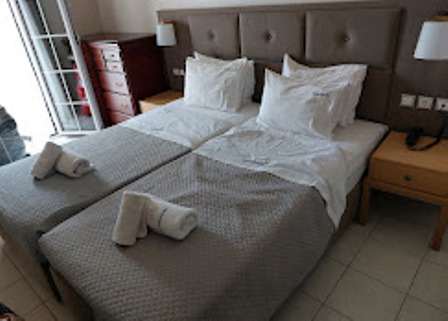 Hotel room with a large double bed, white pillows, gray bed runners, and two nightstands with lamps on either side.