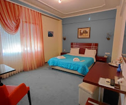 Hotel room with blue and beige decor, queen bed, turquoise bedding, and towels folded on top; orange sheer curtains and a desk on the right.