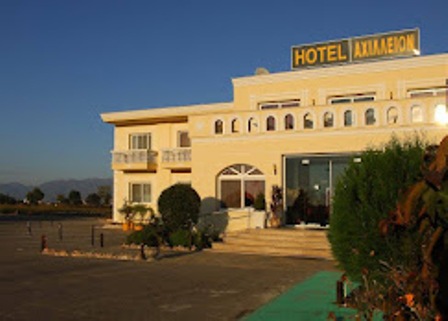 Exterior of a pale-yellow hotel with a HOTEL sign on the roof, glass entrance doors, and landscaped shrubs nearby.