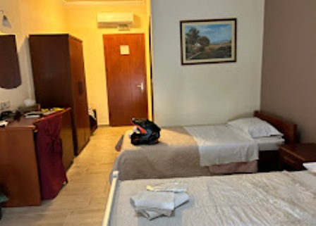 Hotel room with two single beds, a wardrobe, desk, and framed landscape painting; a backpack rests on one bed.
