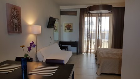 Hotel room with a desk and vase of flowers, a white sofa, wall-mounted TV, a bed, and a balcony door with sheer curtains.