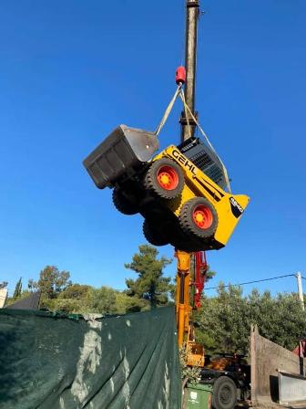 Crane hoists a yellow construction vehicle with large red wheels into the air on a clear day at a construction site.