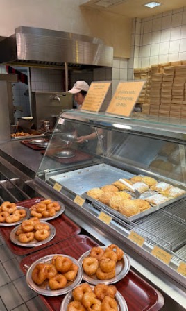 Bakery display case with donuts and pastries; staff member in a white cap behind the counter.