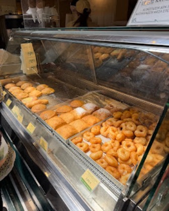 Glass bakery display case filled with assorted donuts and pastries in warm lighting behind a glass barrier appraised for customers